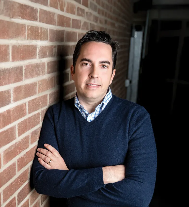Man leaning against brick wall, arms crossed, wearing navy sweater over checkered collared shirt, neutral expression.
