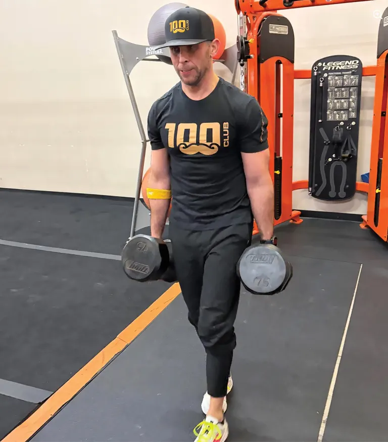 Man in a black 100 Club shirt and cap holding a heavy dumbbell in each hand in a gym