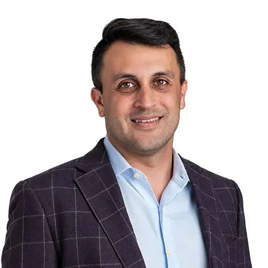 A man in a dark checkered blazer and light blue shirt smiles at camera against white background.