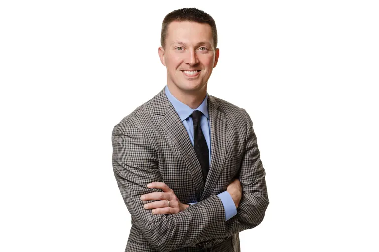 Man in gray checkered suit, blue shirt, tie, arms crossed, smiling against white background.