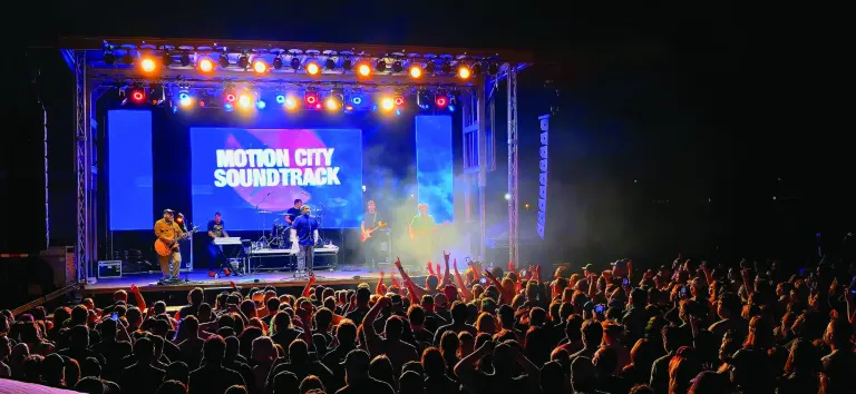 Band playing on a lit outdoor stage under colored lights in front of a large crowd