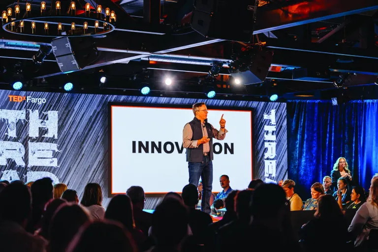 Man in a vest and plaid shirt speaking on a stage at TEDxFargo with a blue curtain and stage lights above