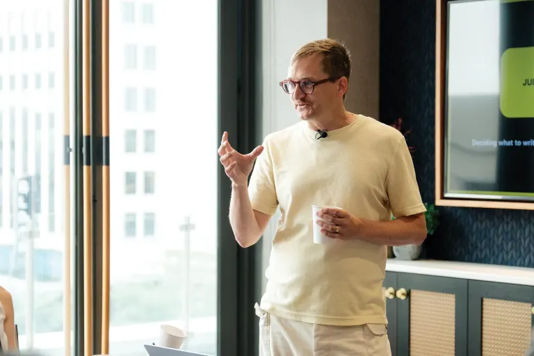 Man wearing beige T-shirt and glasses speaks, gesturing with one hand while holding a cup indoors.