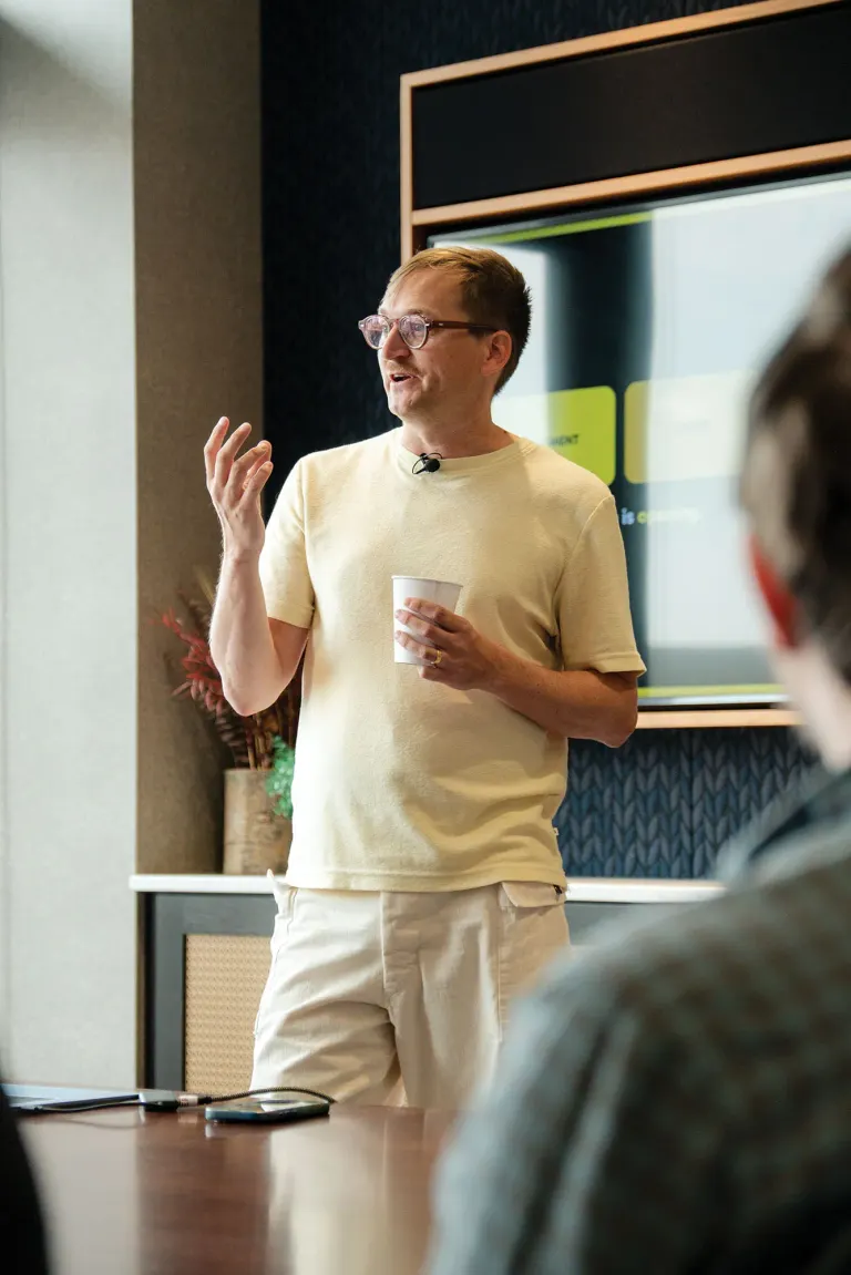 Man in beige t-shirt and glasses speaks, holding a cup, gesturing with one hand, microphone clipped.