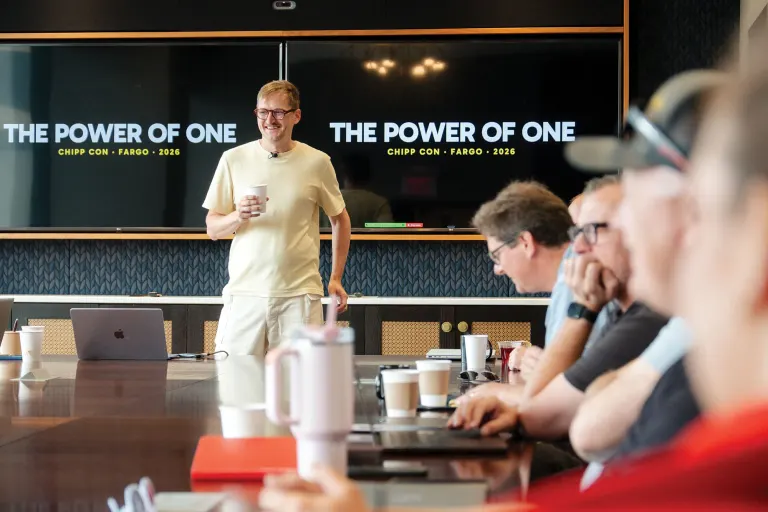 Man standing at front presenting, "THE POWER OF ONE" slides on screen; audience seated around table with laptops.