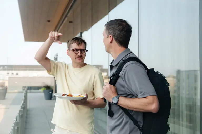 Two men talking on a balcony; one holds a plate of food, the other wears a backpack and gray shirt.