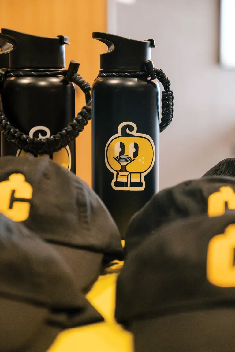 Two black water bottles with yellow cartoon sticker, surrounded by caps with matching logo on yellow.