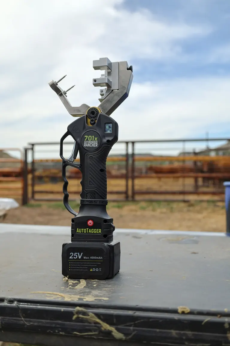 Autotagger device with battery labeled 25V on a metal table, rodeo-style metal adapter top, outdoor pen arena background.