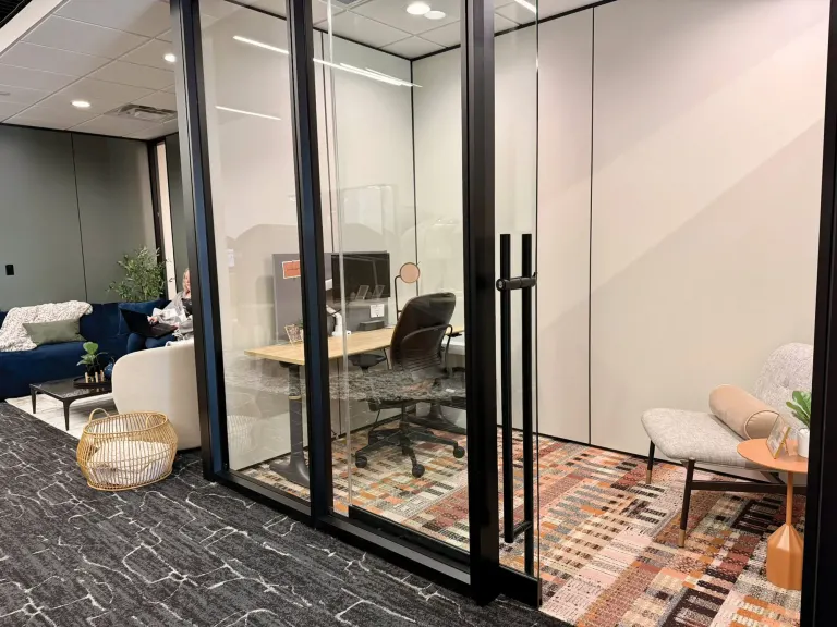Glass-walled private office with a wood desk and task chair, a lounge chair and woven basket in the foreground