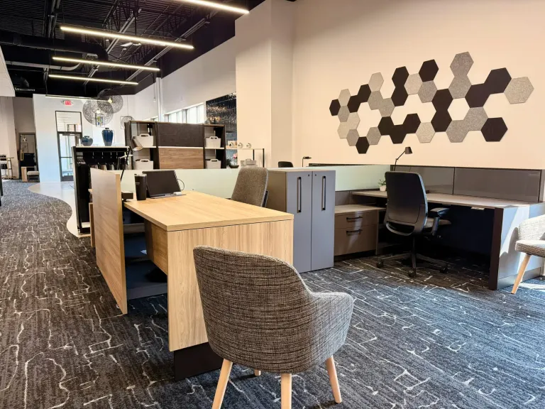 Wood-topped desk with a gray upholstered guest chair, a hexagon felt wall display and workstations behind