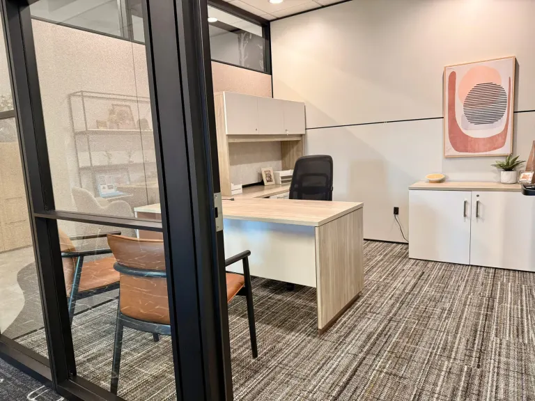 Private office seen through a glass wall, with a light wood L-shaped desk, black chair and white storage cabinet