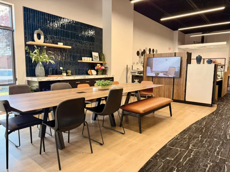 Long wood table with black chairs and a leather bench in front of a dark blue tile wall and a wall-mounted screen