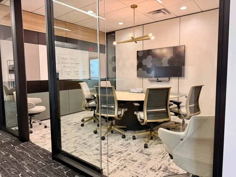 Glass-walled conference room with a round table, rolling chairs, a wall-mounted screen and a whiteboard