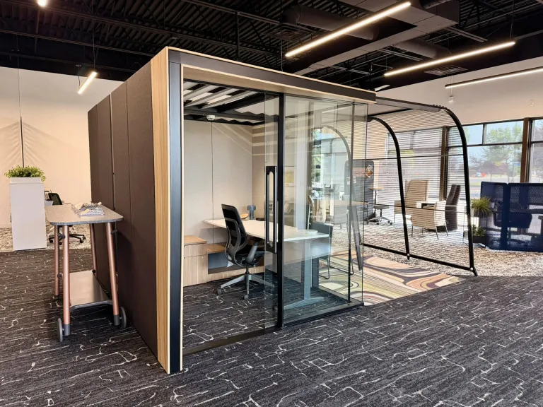 Enclosed glass and wood workpod with a desk and chair inside, set in an open office with black ceiling and linear lighting