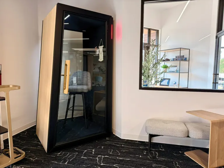 Tall black and wood phone booth next to a window, with a cushioned bench and a standing-height table