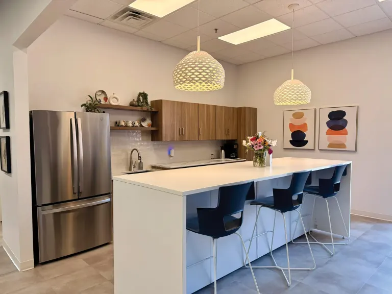 Kitchen area with wood cabinets, a stainless fridge, a white island with dark stools and two woven pendant lights