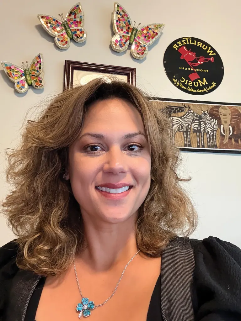 Smiling woman with wavy hair wearing a blue flower pendant necklace; decorative butterflies and wall art behind.