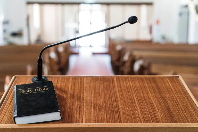 Pulpit with Holy Bible, microphone, and church pews in background, aisle lit by natural light.