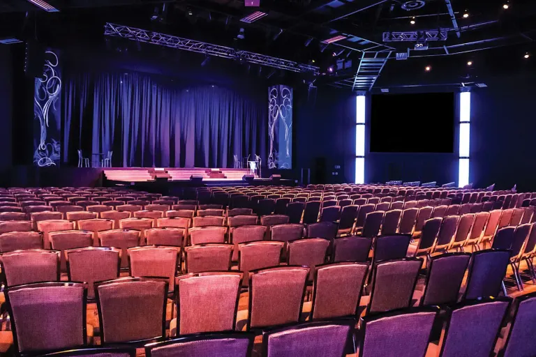 The empty theater with red seats facing a stage and dark curtains under blue lighting.