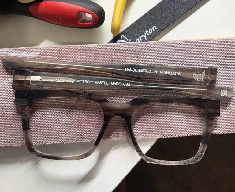Clear rectangular eyeglasses on fabric strip with crafting tools in background; visible text on temple.