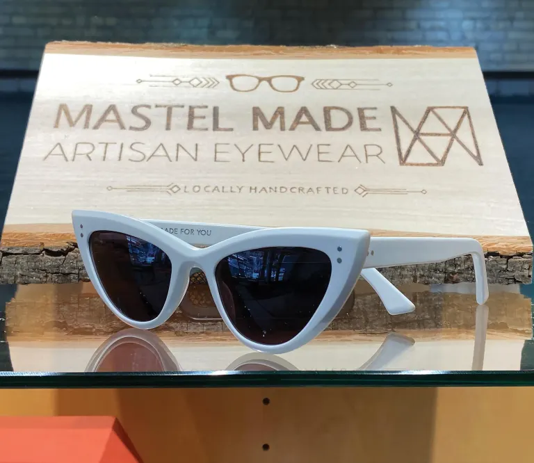 White sunglasses with dark lenses displayed on glass shelf, in front of a wooden “MASTEL MADE ARTISAN EYEWEAR” sign.