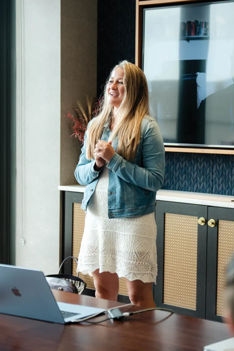 Woman stands speaking in a room, wearing white lace dress and denim jacket, hands clasped, smiling.