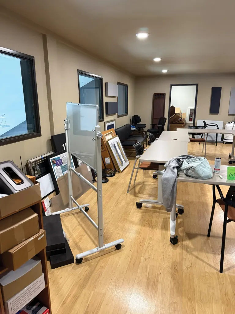 Open room with a rolling whiteboard, folding tables, boxes and framed pictures stacked along the wall