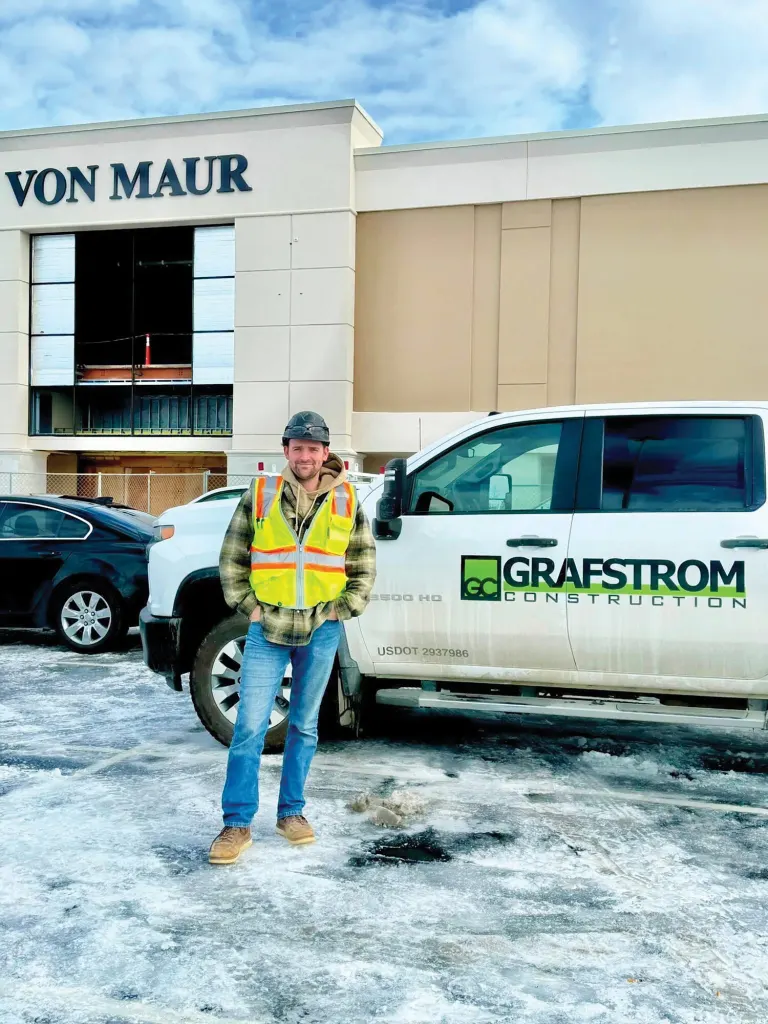 Man in a safety vest standing beside a white Grafstrom Construction truck outside a Von Maur building