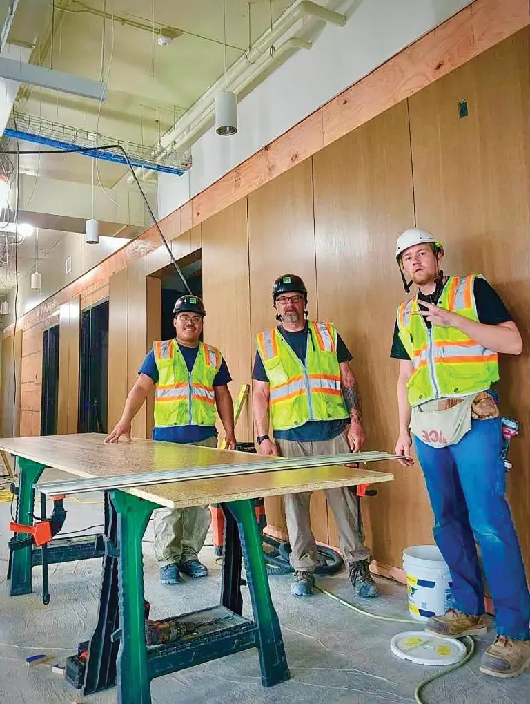 Three workers in safety vests standing around a sawhorse table in a space with wood-paneled walls