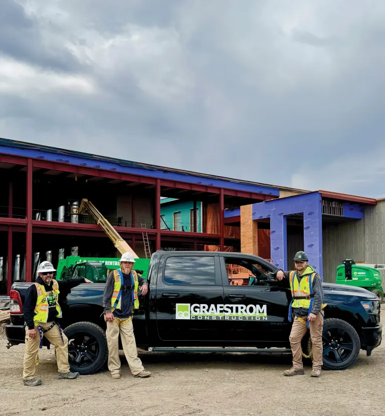 Three workers in hard hats and safety vests standing next to a black Grafstrom Construction pickup truck at a building site