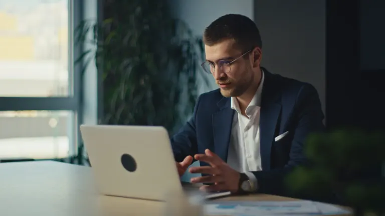 Man in a blazer and glasses working on a laptop at a table by a window