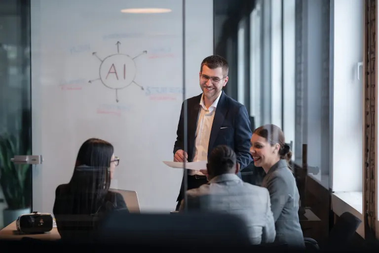 Man standing and smiling at a whiteboard marked AI while a small team sits around a table