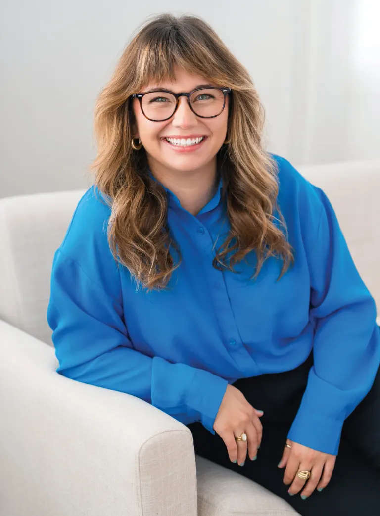 Portrait of Ashley Blazek, smiling, wearing glasses and a blue top, seated on a cream sofa