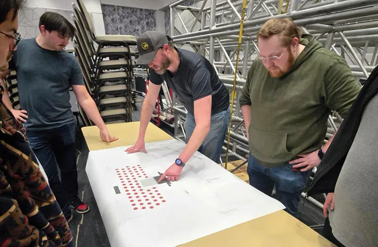 Group of men examining a large blueprint with red dots on a table in a workshop.
