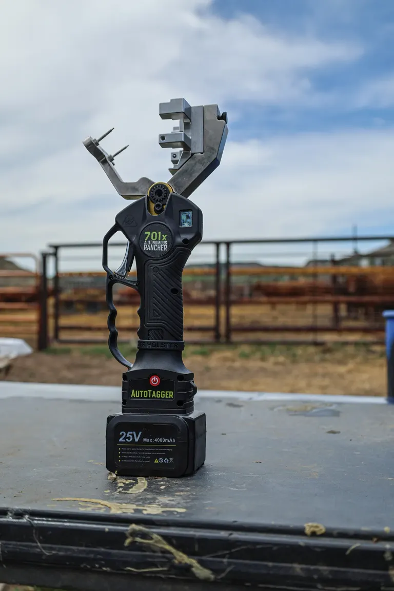 Autotagger device with 25V battery sits on table, metal farming tool attachment above, outdoor farm setting behind.
