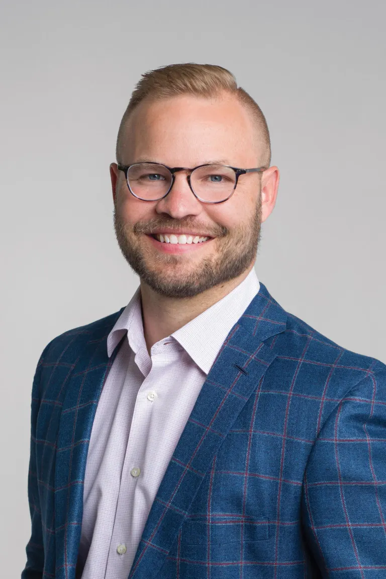 Ben Meland smiling in glasses and a blue plaid blazer against a gray background.