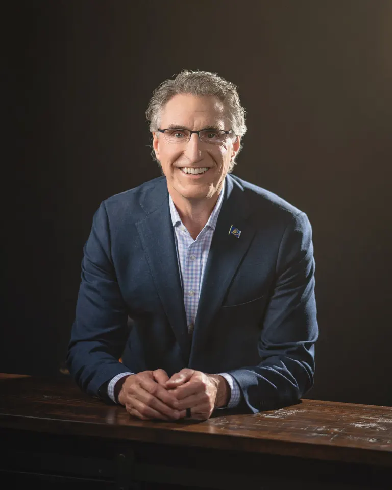 Doug Burgum seated at a wooden table in a navy blazer with his hands clasped, against a dark background.