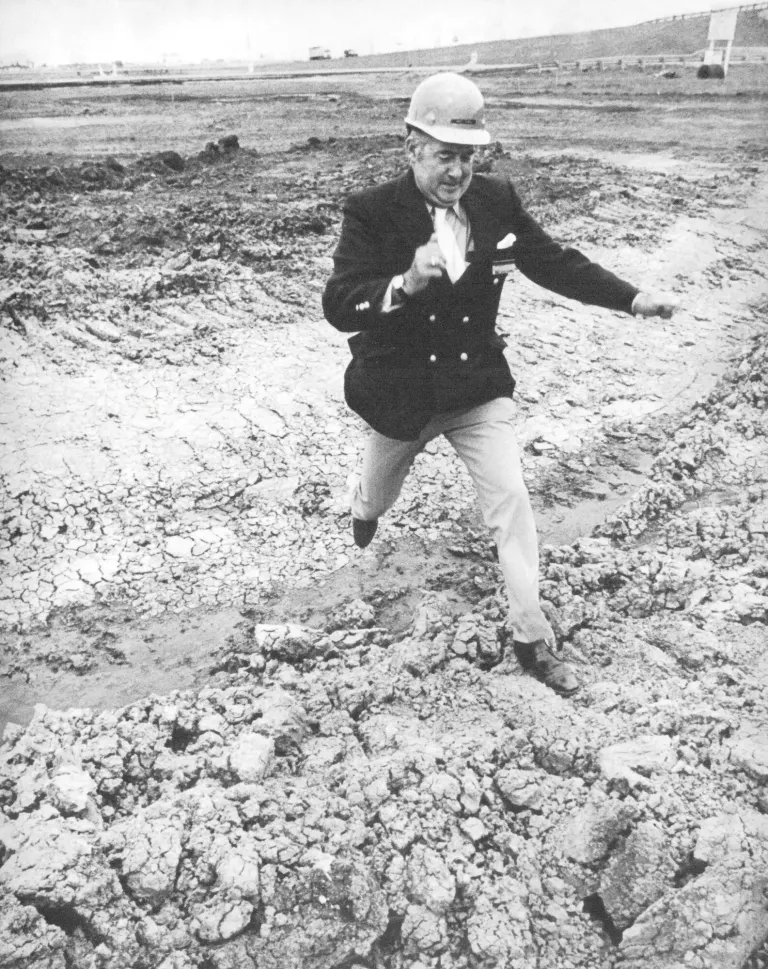 Black-and-white photo of Bill Schlossman in a hard hat and blazer stepping across a muddy construction site.