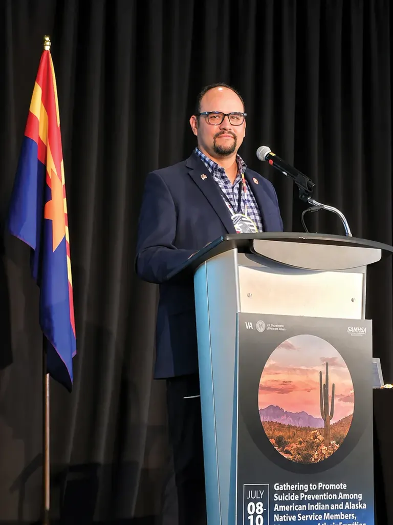 Speaker in dark blazer at podium with microphone, Arizona flag beside stage banner.