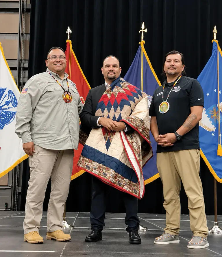 Three men on stage; man in center wrapped in colorful quilt, others in casual uniforms, flags in background.