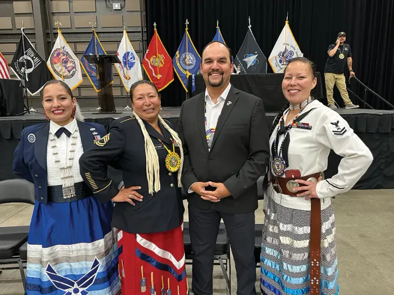 Four people in formal and traditional attire pose together on stage with flags in background.