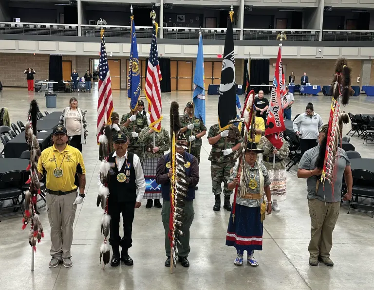 Flag bearers and veterans in ceremonial outfits standing in a hall with flags and medals.