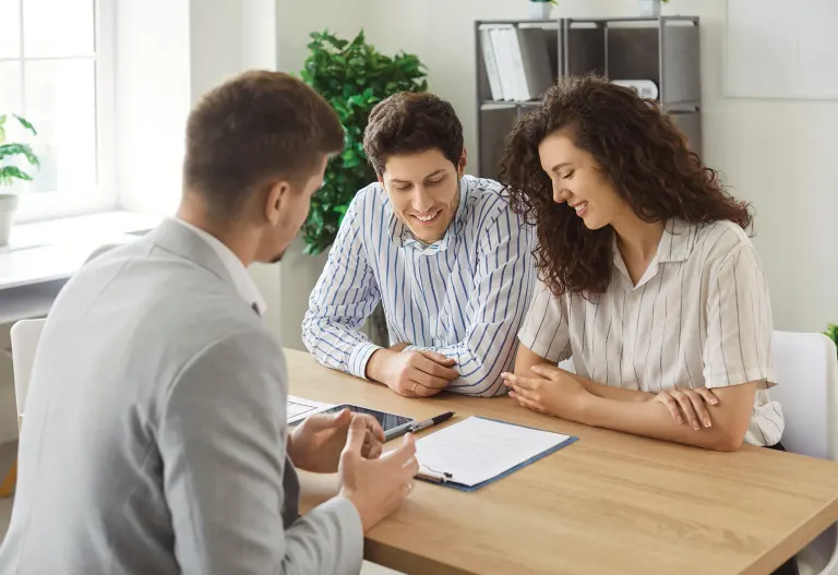 Three colleagues discuss documents at a table in a bright office, smiling and leaning toward each other.