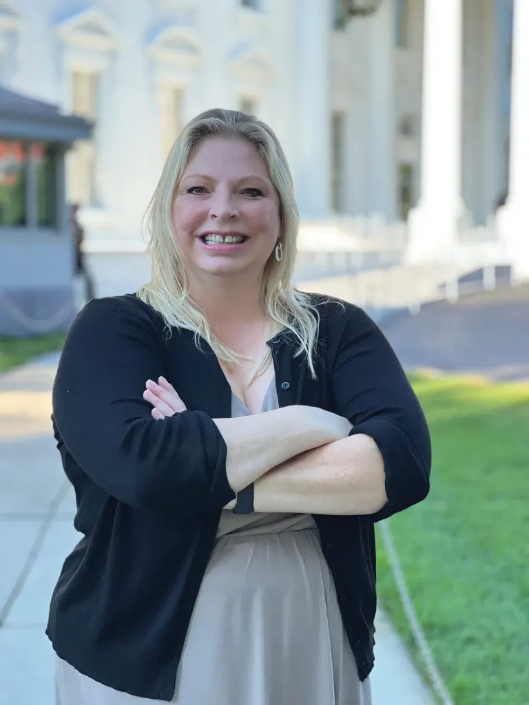 Smiling woman with blond hair and a black cardigan standing with arms crossed outside a white columned building